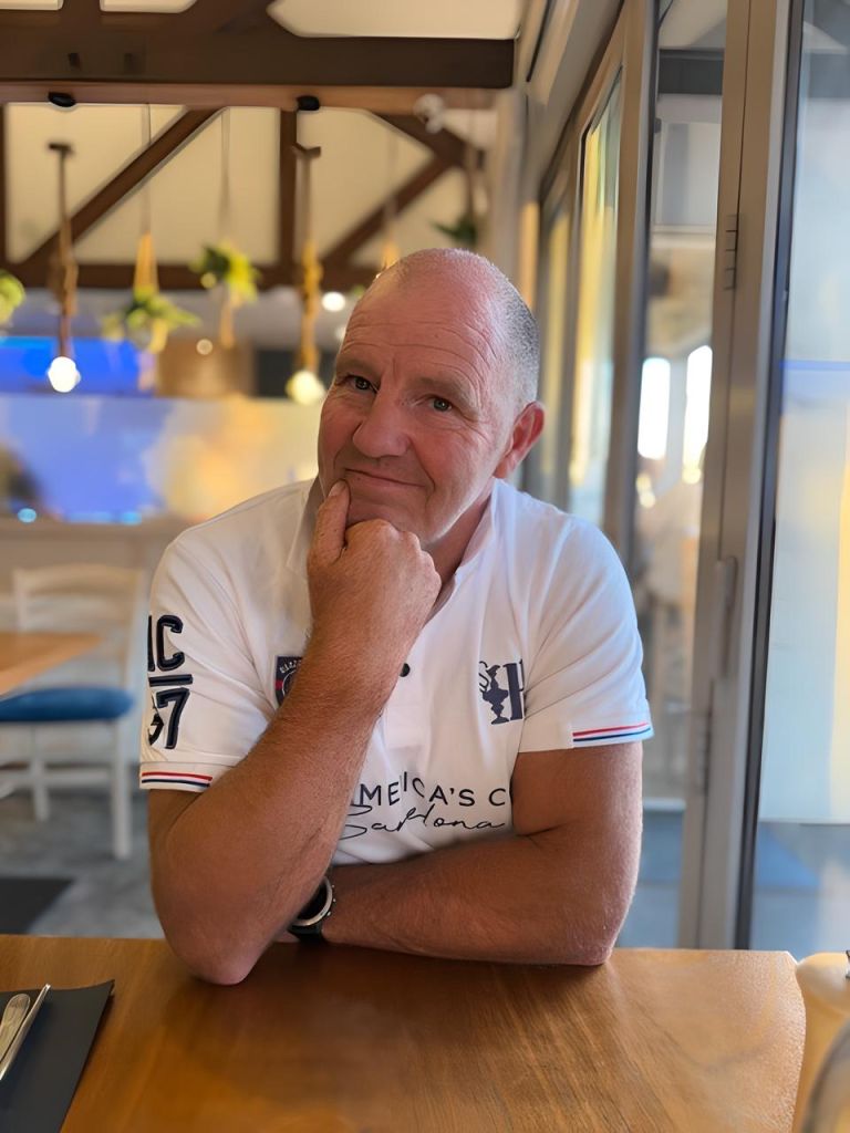 Owner of Wholesale Roofing sitting at a restaurant table, smiling with his hand on his chin, with warm lighting and wooden beams in the background.